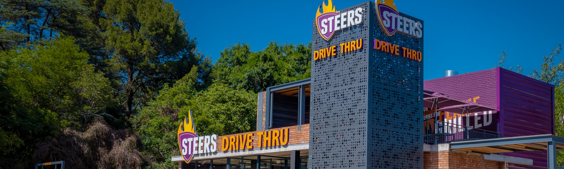 Steers Drive Thru restaurant exterior with branded signage tower surrounded by trees on a sunny day
