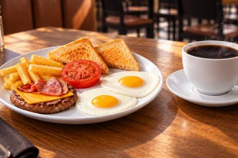 Hearty sit-down breakfast with fried eggs, beef patty, bacon, cheese, chips, tomato, toast and coffee