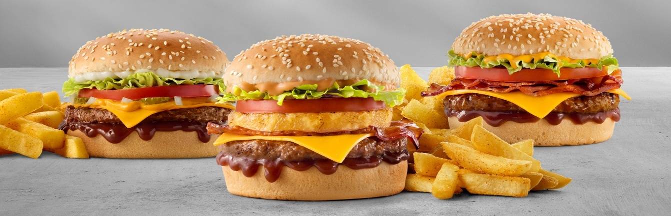 Three classic cheeseburgers with crispy bacon, onion rings, lettuce, tomato, melted cheddar cheese, barbecue sauce, sesame seed buns, and chips