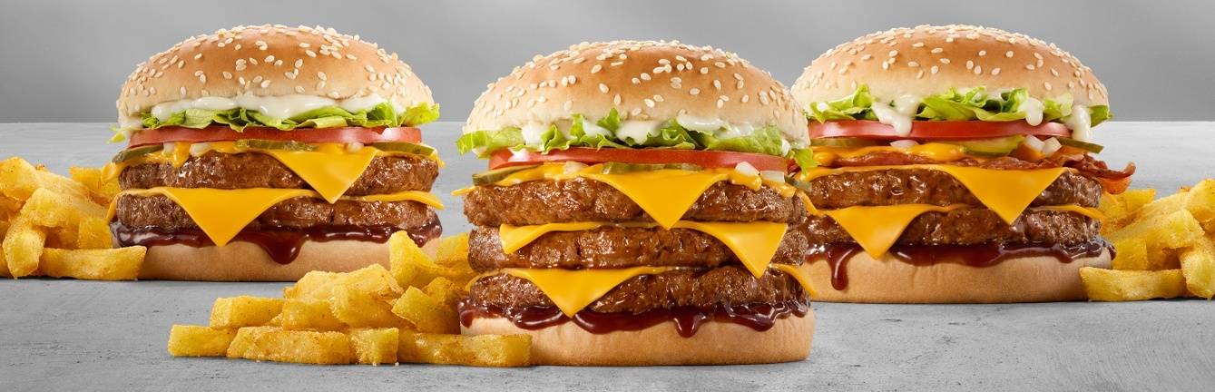 Three stacked cheeseburgers with double and triple beef patties, melted cheddar cheese, lettuce, tomato, pickles, sesame seed buns, barbecue sauce, and fries