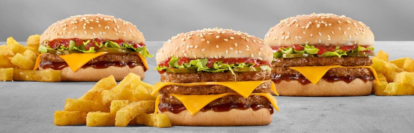 Three stacked double cheeseburgers with melted cheddar cheese, lettuce, pickles, ketchup, sesame seed buns, and crispy French fries on a gray background