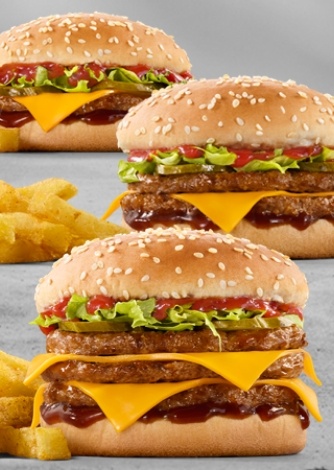 Three stacked double cheeseburgers with melted cheddar cheese, lettuce, pickles, ketchup, sesame seed buns, and crispy French fries on a gray background
